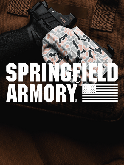 A handgun resting on a brown tactical vest, with a camouflage-patterned holster covering the grip. Large white text across the center reads 'SPRINGFIELD ARMORY' with a U.