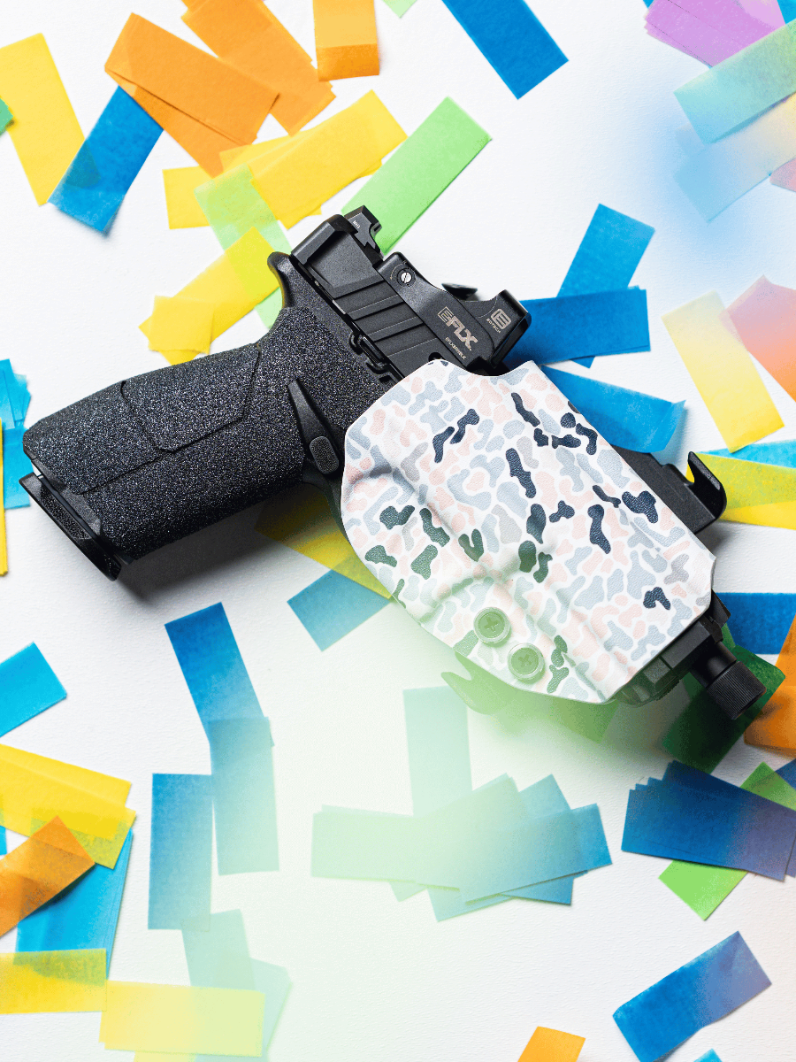 A black semi-automatic handgun resting on a white surface scattered with colorful paper confetti. The gun is partially covered by a white holster with a green and gray camouflage pattern.